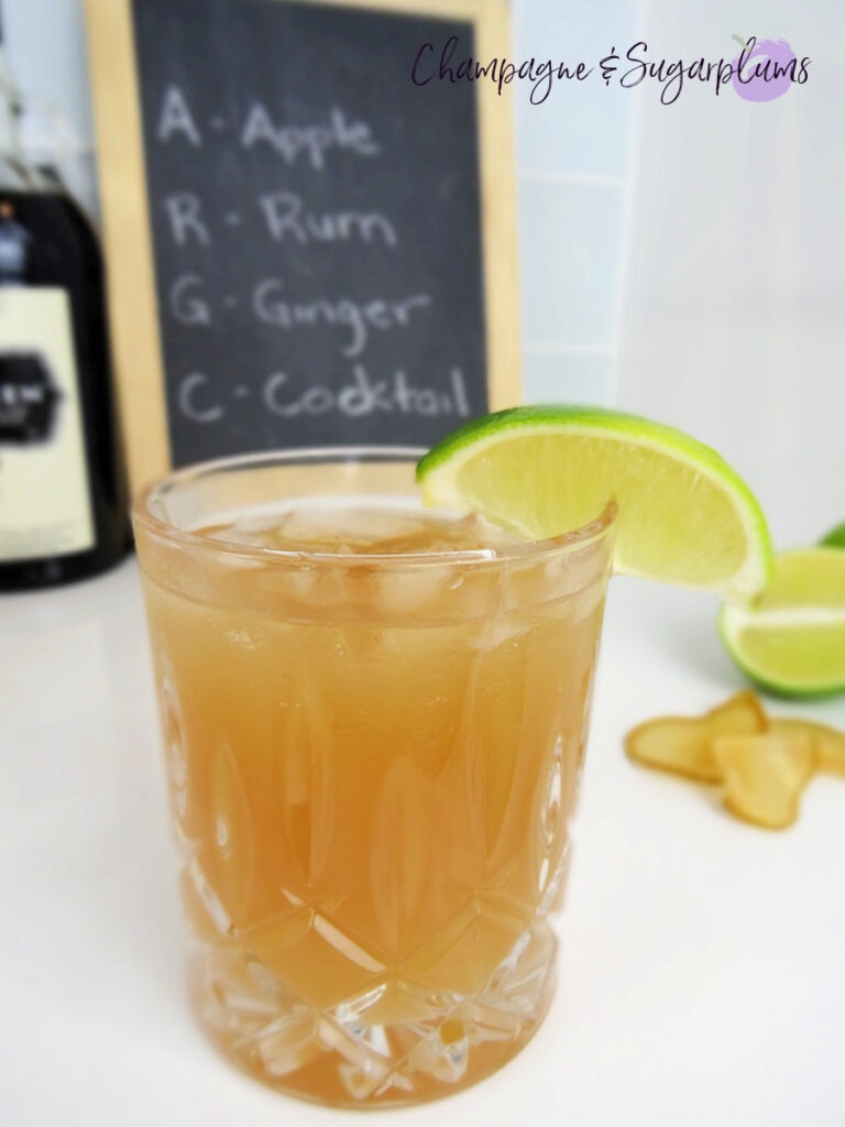 The Teacher's Pet Back to School Cocktail in a highball glass, garnished with a lime wedge on a white background with limes and chalkboard by Champagne and Sugarplums