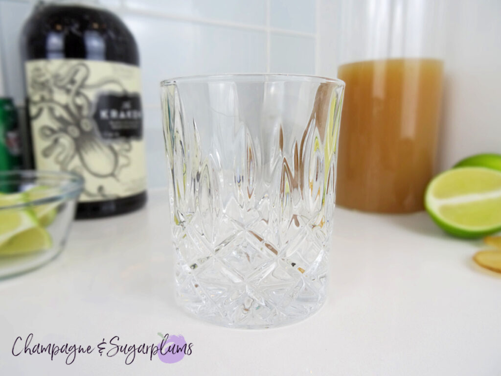 A highball glass, rum bottle, and apple cider in a glass carafe on a white background with limes by Champagne and Sugarplums