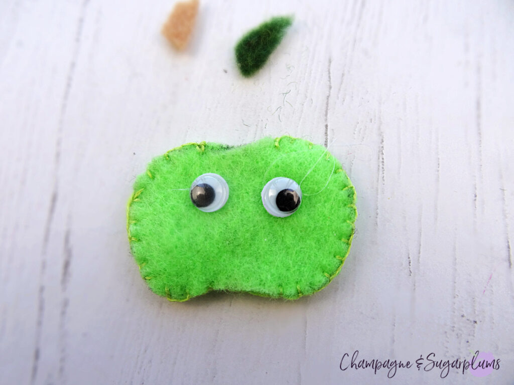 Gluing google eyes onto a felt green apple on a white background by Champagne and Sugarplums