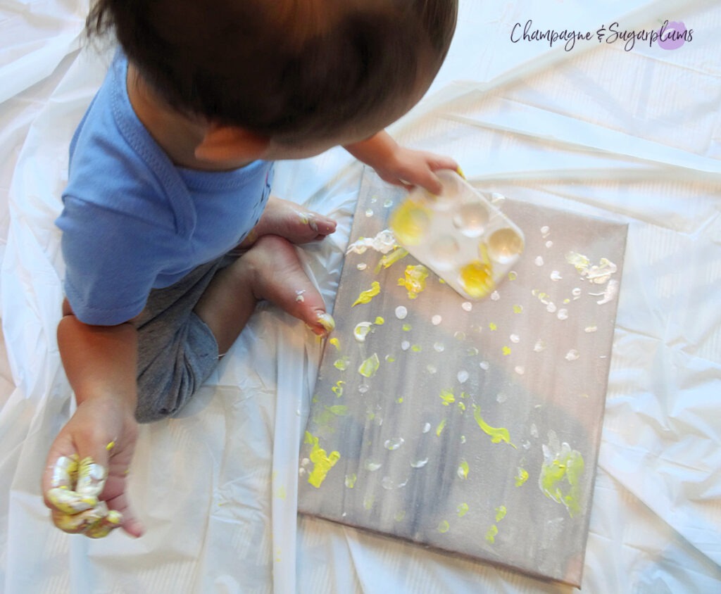 A toddler painting white and yellow dots onto a gray canvas by Champagne and Sugarplums